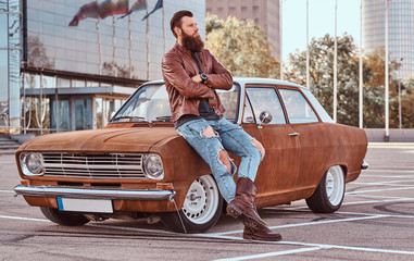Bearded male dressed in brown leather jacket and boots leaning on tuned retro car in the city parking near skyscraper.
