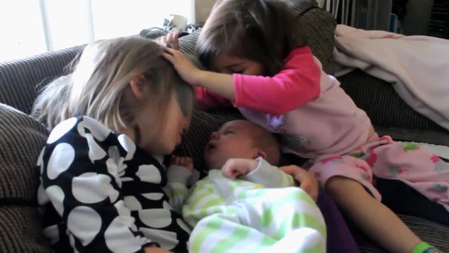 Two sisters fight over who gets to kiss newborn baby brother.  One is holding and kissing him, the other pushes her head back and leans in to kiss him herself.  Home video of kids.