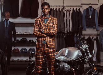 Fototapeta na wymiar African-American man dressed in a trendy elegant suit posing with crossed arms near retro sports motorbike at the men's clothing store.
