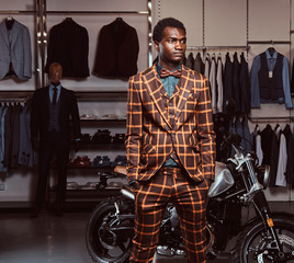 African-American man dressed in a trendy elegant suit posing with hands in pockets near retro sports motorbike at the men's clothing store.