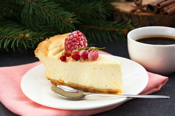 Cheesecake, decorated frozen raspberry and red currant berries, and coffee cup on pine tree branch background. Christmas dessert with summer taste