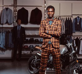 Fototapeta na wymiar African-American man dressed in a trendy elegant suit posing with crossed arms near retro sports motorbike at the men's clothing store.