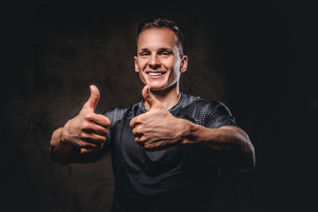 Smiling young sportsman showing thumbs up on dark background.