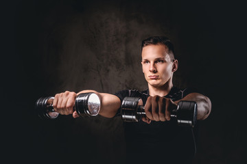 Naklejka na ściany i meble A young bodybuilder wearing sportswear doing exercise with dumbbells on dark background.