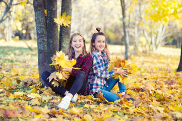 Two beautiful model teenage girls sitting in golden foliage in colorful park, laugh and drop yellow leafs