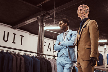 Elegantly dressed African-American man posing with crossed arms near mannequin in a classic menswear store.