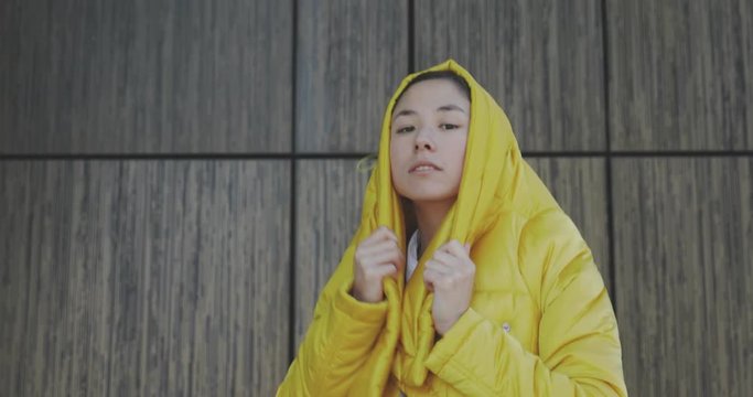 Outdoors fashion portrait of trendy pretty asian girl posing on the wall background. Woman smiling and walking on the city. Wearing stylish wide jeans and long yellow jacket. 4k video