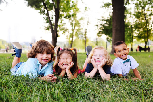 Four Friends Of Preschool Kids Playing Lying On The Grass In The Park On A Summer Day