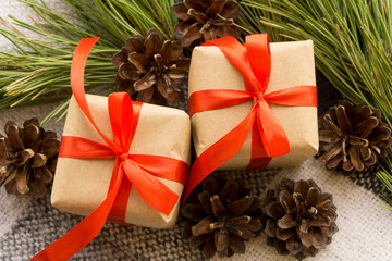 A couple of Christmas gifts in craft paper, with red bows, lies in the middle of pine branches and pine cones.