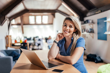 Portrait of a smiling blonde woman looking at camera and using laptop.