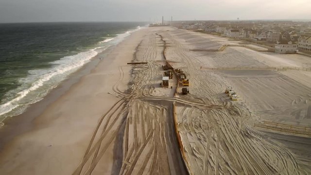 Fast fly by aerial pull back of the large scale dredging operation of a long pipe laying along the beach spraying new sand and construction equipment redirecting it.
