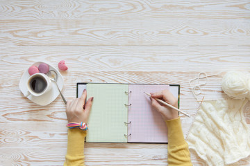 A woman knits a white knitted fabric of woolen yarn. Coffee black. Pastry macaroons. An old background of pastel color.