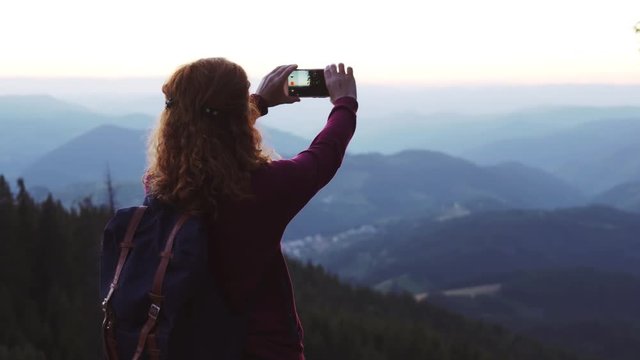 Slow motion a slide young red-haired woman stands on top of a high mountain against the background of a large mountain lake and takes a selfie on a smartphone on the sunset day. Relax. Lens flare