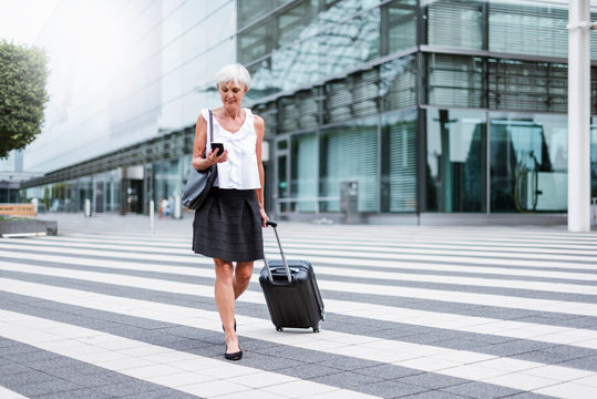 Smiling senior woman with baggage on the move looking on cell phone