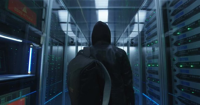 Medium, slow motion shot from behind of a hacker with a backpack walking through corporate data center with rows of working rack servers