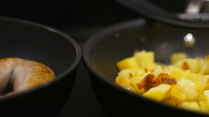 Sausage and potatoes being fried on pans on a glasshob in a kitchen with a pan left to right.