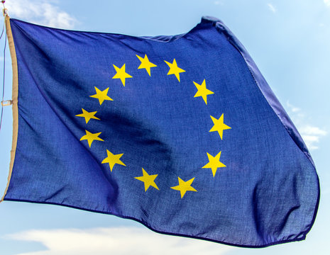 Flag Of The European Union Flutters On The Blue Sky. A Blue Flag With Yellow Stars In Circles - The EU Symbol Winds In The Wind.