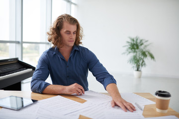 Young musicican with long hair looking at music sheets on his table in studio