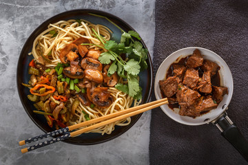 Chinese noodles with mushrooms, fried meat in a pan, vegetables and herbs are on the gray table. Top view