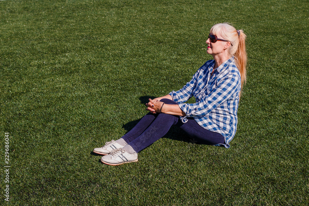 Wall mural well-groomed caucasian woman fifty years in a plaid shirt and jeans sitting on the grass