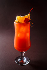 Glass of red alcohol cocktail with cherry, slice of orange and straw on elegant dark brown background