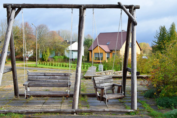 Fototapeta na wymiar Empty natural wooden swing with two sits in yard with residential buildings and trees on background in rainy autumn day.