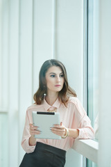 Fototapeta premium Pensive young female entrepreneur with tablet computer standing at office window