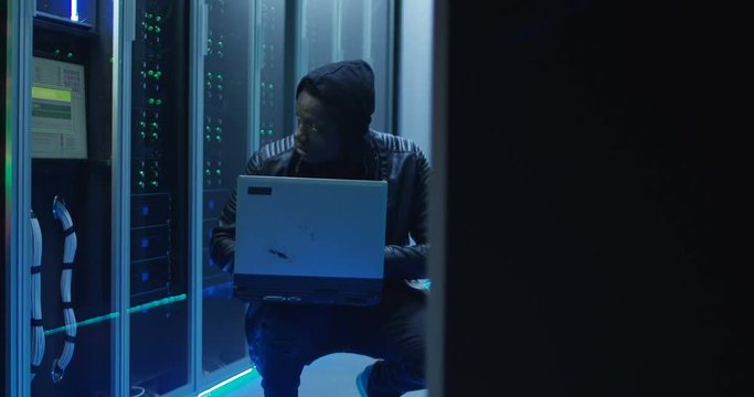 Medium shot of a hacker in a hoodie on laptop in corporate data center with rows of working rack servers