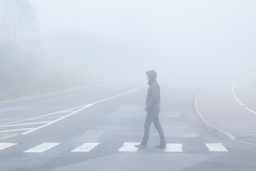 Young man alone without reflector elements crossing street on crosswalk in mist of early morning....