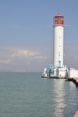 Fototapeta na wymiar Lighthouse of Odessa port in Ukraine. Old white beacon with reflection in calm sea water surface and seascape background. Red lantern lamp on the top of lighthouse building. Black sea view