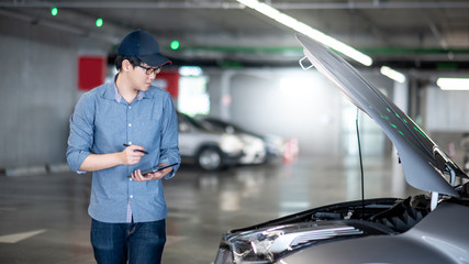 Asian auto mechanic holding digital tablet checking car engine under the hood in auto service garage. Mechanical maintenance engineer working in automotive industry. Automobile servicing and repair