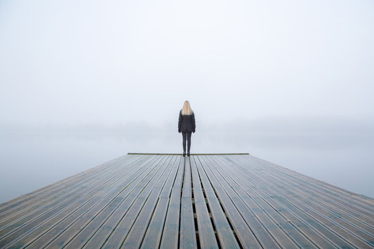 Young woman standing alone on edge of footbridge and staring at lake. Mist over water. Foggy air. Early chilly morning in autumn. Beautiful freedom moment and peaceful atmosphere in nature. Back view.