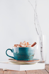 Hot chocolate with marshmallows and cinnamon stick in a blue ceramic cup on a table with a book. The concept of winter or fall time. Minimal scandinavian design.