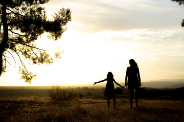 Fototapeta na wymiar Backlight of a mother and her daughter walking holding hands in a pine forest with yellow sky