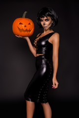 Sexy brunette woman in Halloween makeup and lingerie hold a pumpkin on a black background in the...