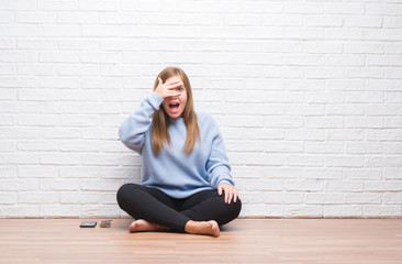 Young adult woman sitting on the floor in autumn over white brick wall peeking in shock covering face and eyes with hand, looking through fingers with embarrassed expression.