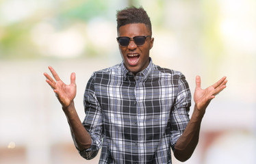 Naklejka premium Young african american man wearing sunglasses over isolated background celebrating crazy and amazed for success with arms raised and open eyes screaming excited. Winner concept