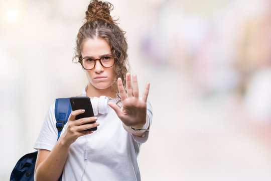 Young brunette student girl wearing backpack, headphones and smartphone over isolated background with open hand doing stop sign with serious and confident expression, defense gesture