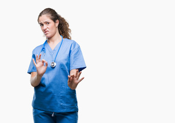 Young brunette doctor girl wearing nurse or surgeon uniform over isolated background disgusted expression, displeased and fearful doing disgust face because aversion reaction. With hands raised