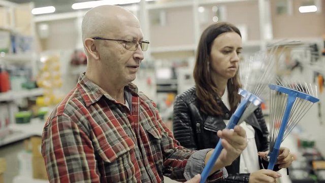 An elderly man with adult daughter on the shelves of the supermarket business choose a garden rake.