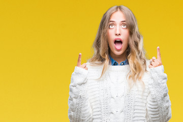 Beautiful young blonde woman wearing winter sweater over isolated background amazed and surprised looking up and pointing with fingers and raised arms.