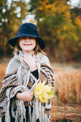 Very emotional autumn portrait of a cute little girl in a field near the forest. Atmospheric autumn background. Little cute girl in a poncho and a hat in a field with several leaves in her hand. 