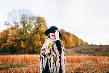 Very emotional autumn portrait of a cute little girl in a field near the forest. Atmospheric autumn background. Little cute girl in a poncho and a hat in a field with several leaves in her hand. 