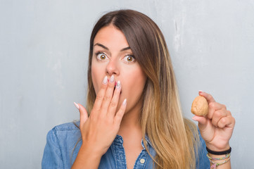 Young adult woman over grey grunge wall showing dried walnut cover mouth with hand shocked with shame for mistake, expression of fear, scared in silence, secret concept