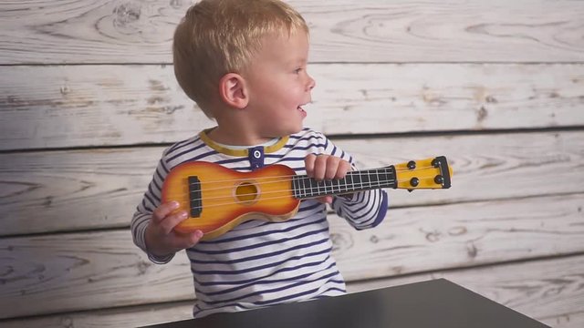 happy one year old boy plays his guitar or ukulele and sings songs, sitting in the wooden room.