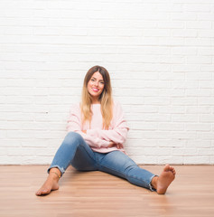 Young adult woman sitting on the floor over white brick wall happy face smiling with crossed arms looking at the camera. Positive person.