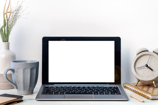 Hipster wooden desk with an open laptop mockup, a bright vase with green grass, a notebook, a notebook, a gray cup of coffee and a concrete clock