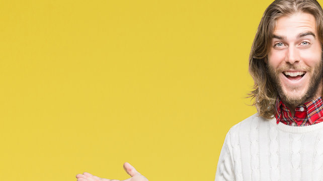 Young handsome man with long hair wearing winter sweater over isolated background amazed and smiling to the camera while presenting with hand and pointing with finger.