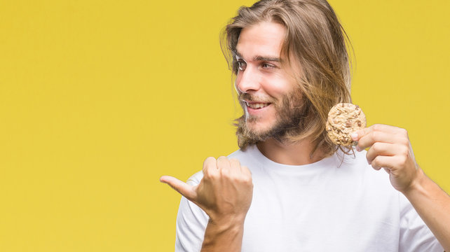 Young handsome man with long hair eating chocolate cooky over isolated background pointing and showing with thumb up to the side with happy face smiling