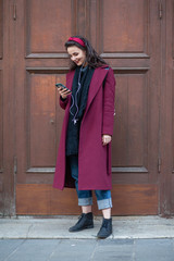 young smiling woman in coat listen music with wire headphones near vintage wooden door on autmun street